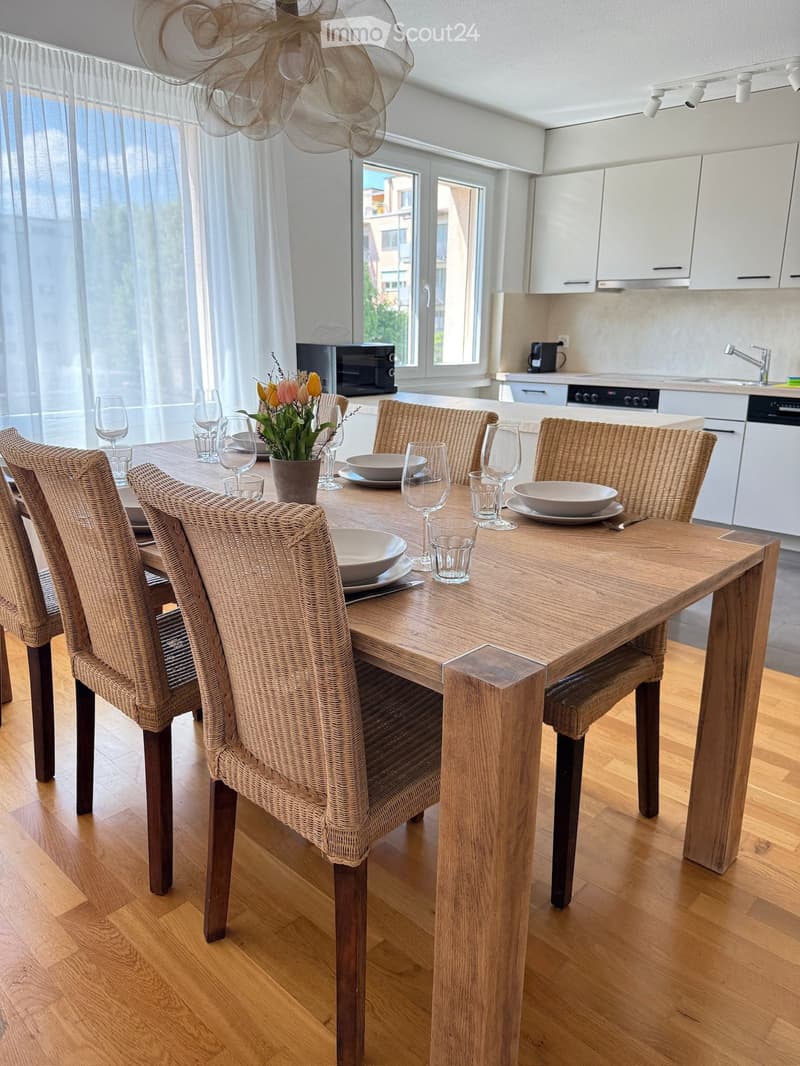 A modern dining area with a wooden dining table surrounded by wicker chairs, set with plates, bowls, and glasses. The area is adjacent to a sleek kitchen with white cabinets, a sink, and various appliances.