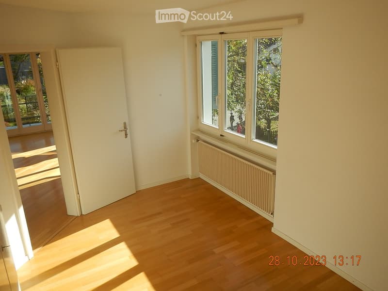 Empty room with light wooden flooring, white walls, and large windows with a radiator below. A white door leads to another space.