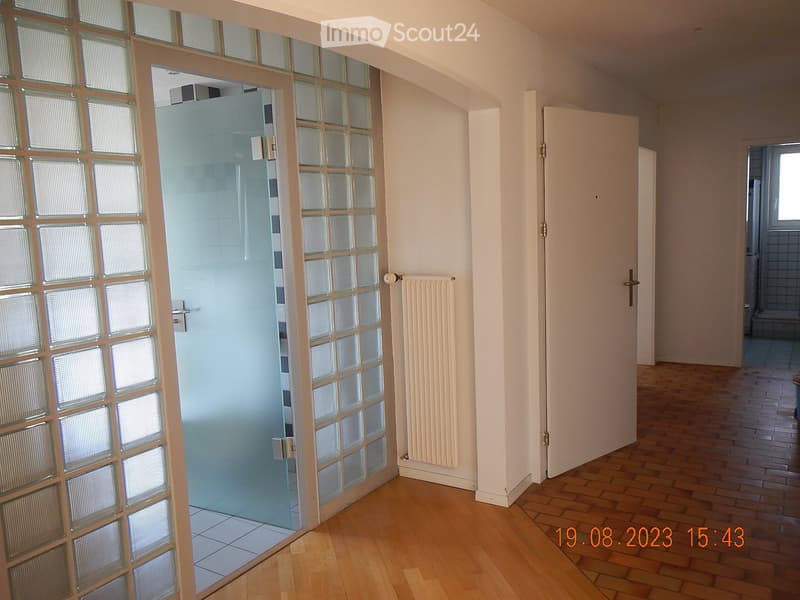 Empty hallway with wooden flooring, white walls, glass-paneled door leading to a tiled room, and a radiator on the wall.