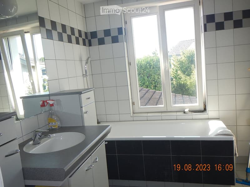 Modern bathroom with white tiles, black accents, a large window, a sink with a faucet, a bathtub, and a cabinet with cleaning supplies.