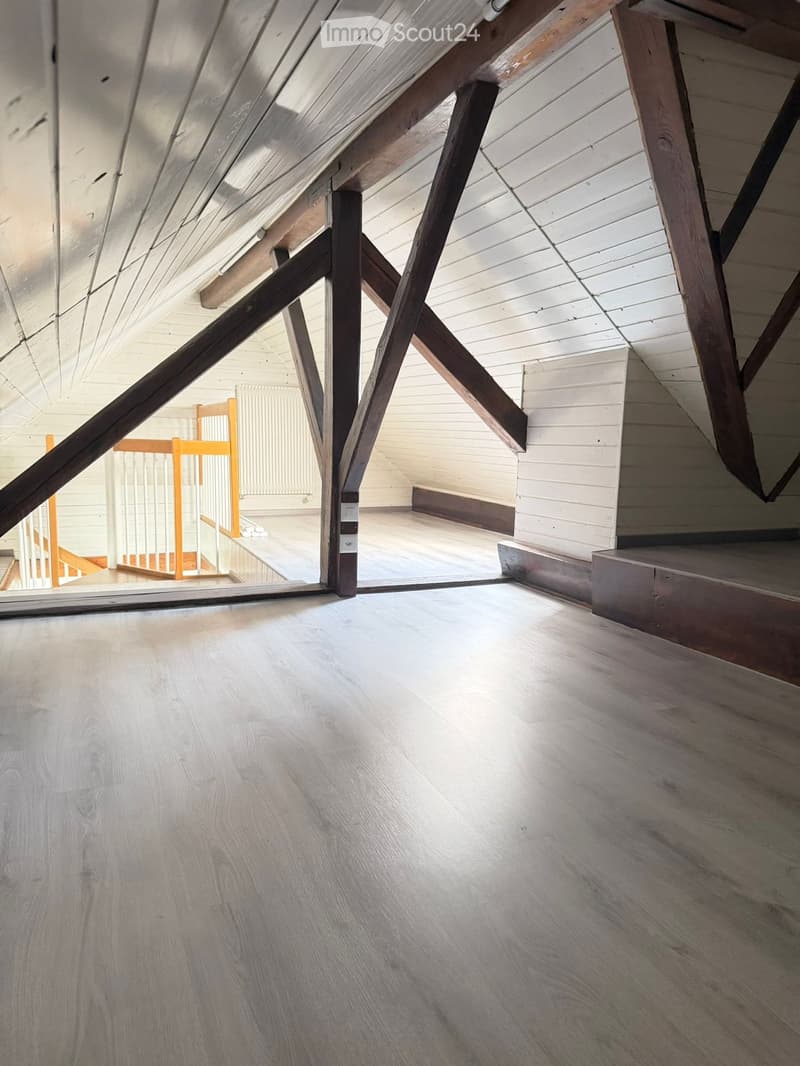 An attic space with exposed wooden beams and a slanted wooden ceiling. The floor is light-colored wood, and there is a small wooden staircase leading to another level.