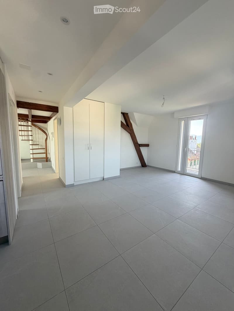 Empty room with tiled floor, white walls, and a sliding glass door leading to a balcony. A white cabinet and a wooden staircase are visible in the background.
