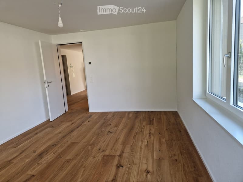 Empty room with wooden flooring, white walls, and a large window with a white sill. An open door leads to another room.