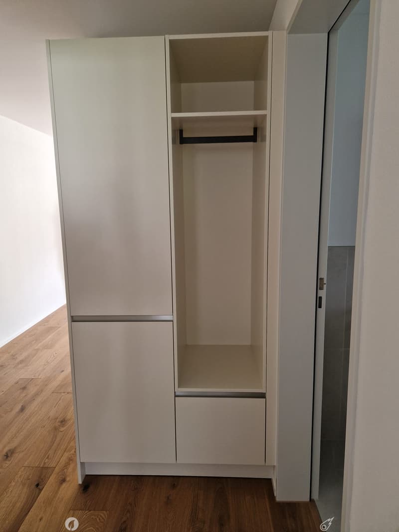 Empty closet with light-colored wooden doors and shelves, wooden floor, white walls.