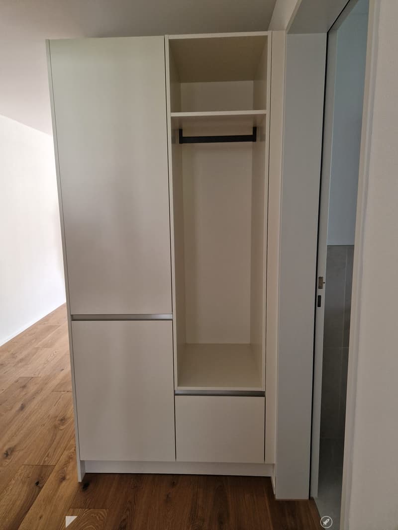 Empty closet with light-colored wooden doors and shelves, wooden floor, white walls.