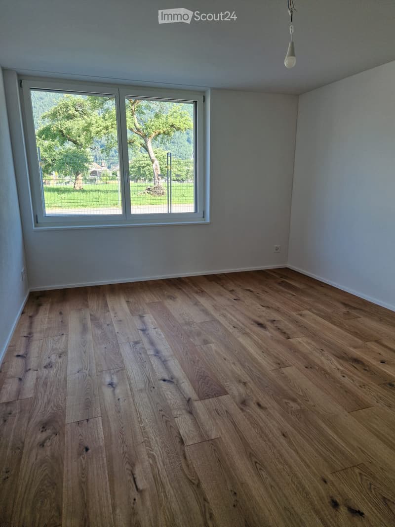 Empty room with light wooden flooring and large windows offering a view of greenery outside.