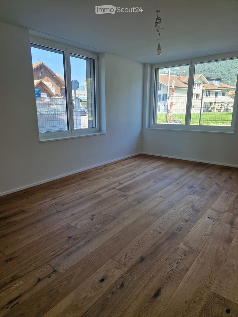 Empty room with wooden flooring and large windows offering a view of the neighborhood.