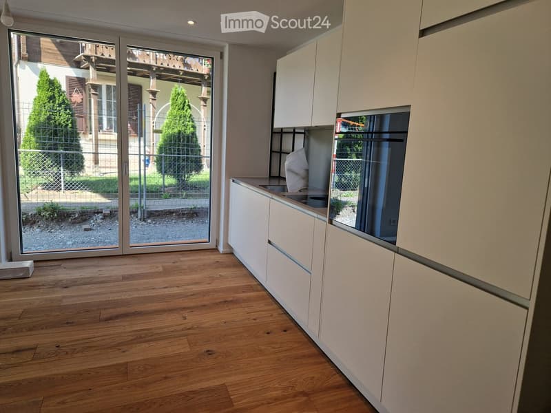 Modern kitchen with light-colored cabinets, wooden flooring, and a large sliding glass door leading to an outdoor area.