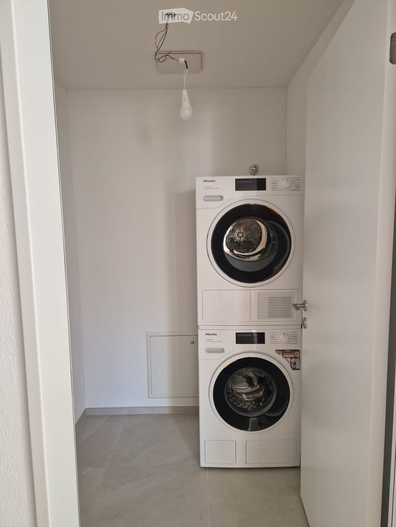 Compact bathroom with a stacked washing machine and dryer unit, white tiled walls, and a single light fixture.