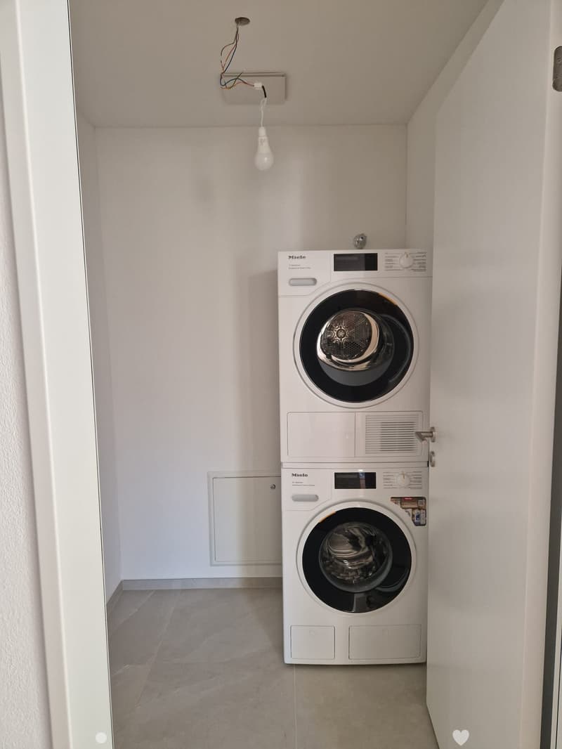 Compact bathroom with a stacked washing machine and dryer unit, white tiled walls, and a single light fixture.