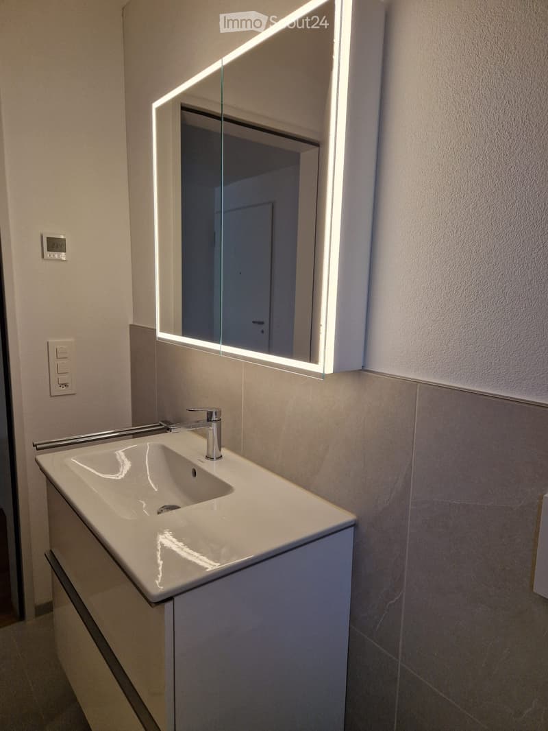 Modern bathroom with a white sink, chrome faucet, and illuminated mirror above it. The walls are tiled in neutral tones, and the space is clean and contemporary.