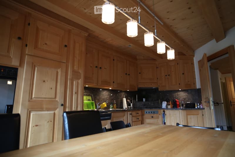 Bright kitchen with wooden cabinets, a central wooden table, and modern appliances including a refrigerator, oven, and microwave. The space features a dark countertop and a hanging light fixture above the table.