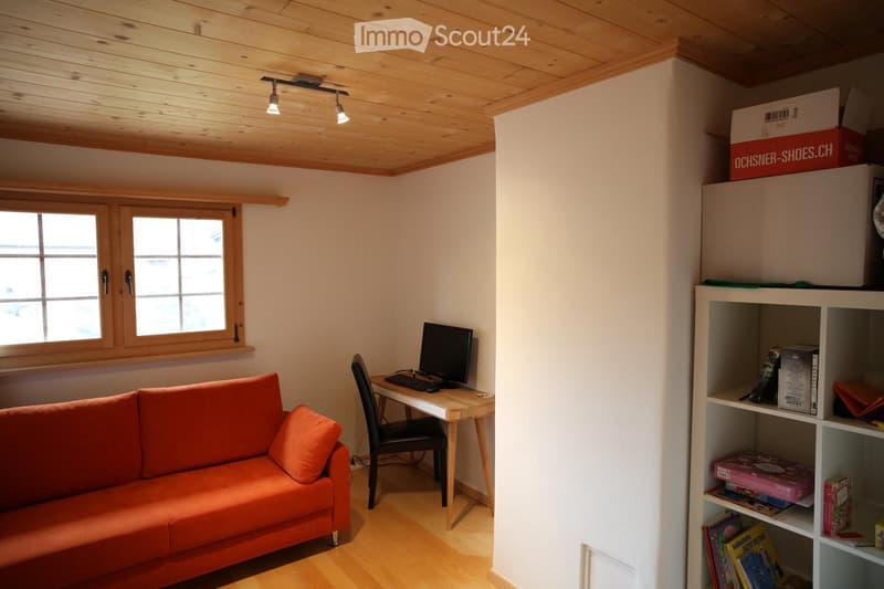 Bright, cozy room with wooden ceiling and floor, large window, orange sofa, desk with computer, and shelving unit with books and boxes.