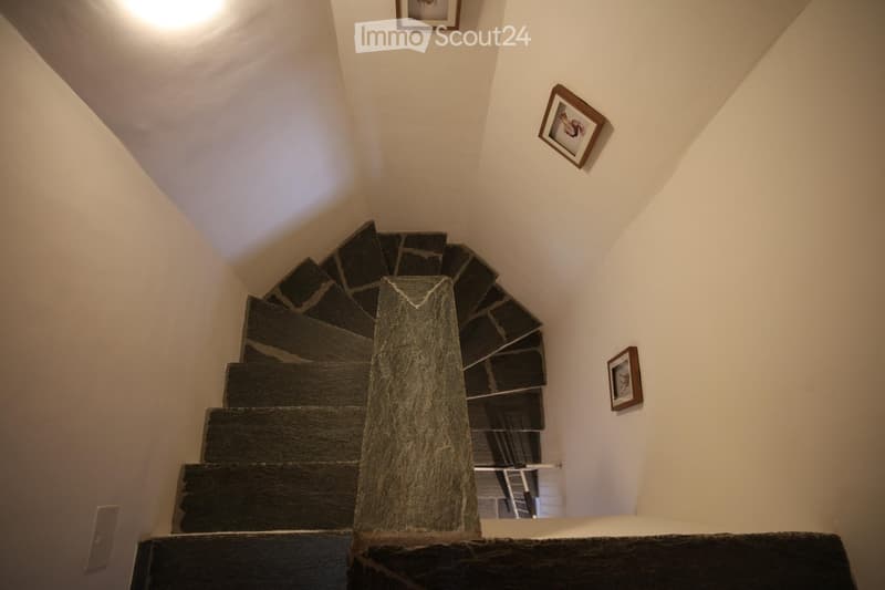 Spiral staircase with dark stone steps and a central wooden handrail, surrounded by white walls and framed artwork.