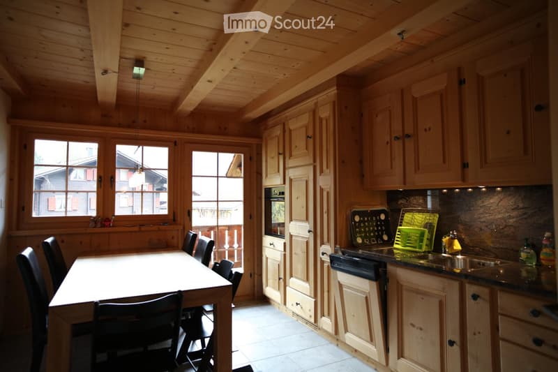 Bright, wooden kitchen with a dining table and chairs, wooden cabinets, a sink, and a dishwasher. Large windows and a door lead to a balcony.