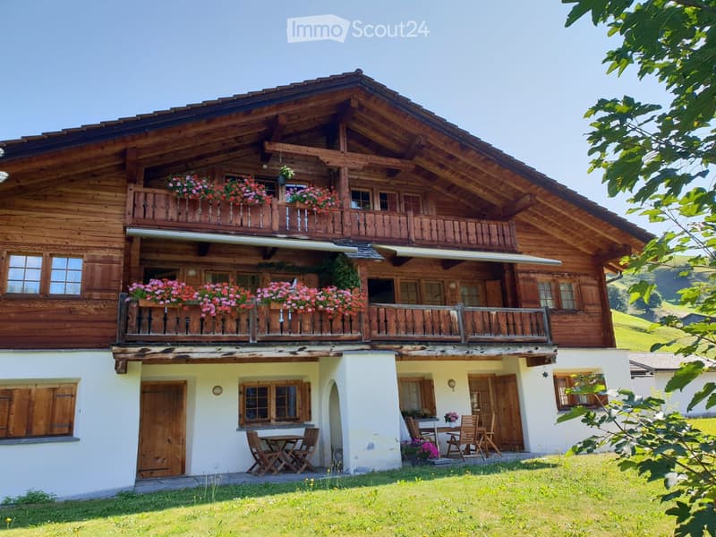 A two-story wooden house with balconies adorned with pink flowers, a well-maintained lawn, and outdoor seating area with tables and chairs.