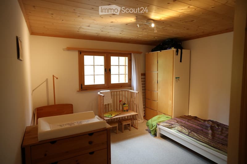 Cozy bedroom with wooden ceiling, large window, bed, baby crib, small table with books, wardrobe, and a green blanket on the floor.