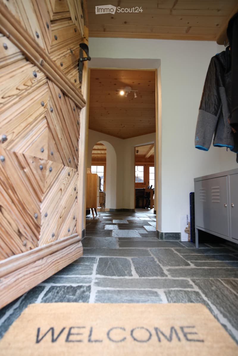 A spacious hallway with wooden walls, a stone-tiled floor, and a coat rack with jackets. The hallway leads to another room with visible furniture and lighting.