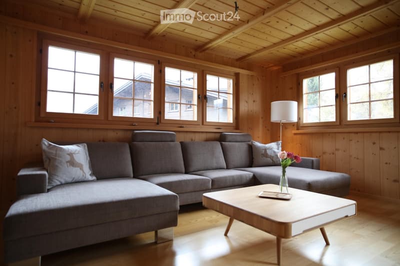 Cozy living room with wooden walls and ceiling, large windows, grey sectional sofa, wooden coffee table, floor lamp, and decorative vase with flowers.