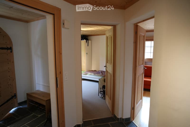 A narrow hallway with wooden doors leading to various rooms, including a bedroom and a living area. The hallway features a wooden bench and a small storage unit on the left side.