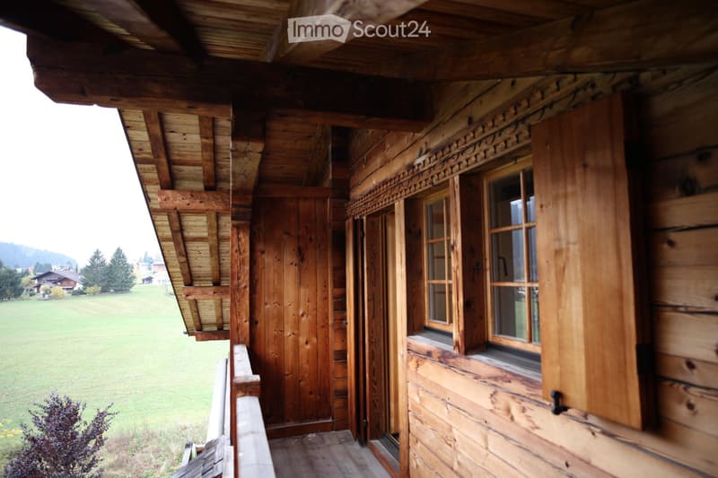Wooden balcony with open wooden railings, large windows, and a scenic view of a green field and distant mountains.