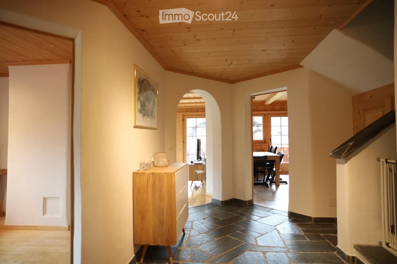 A well-lit hallway with wooden ceiling beams, stone flooring, and a small wooden console table with decorative items. The hallway features arched doorways leading to other rooms, including a dining area visible in the background.