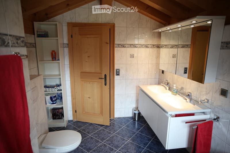 Bathroom with a white sink, mirror, toilet, wooden door, and shelves holding various items. The room features tiled walls and a slanted wooden ceiling.