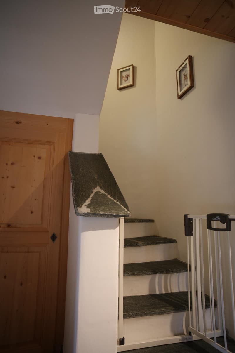 A narrow staircase with wooden steps and a white railing, featuring a small rug on the landing and framed pictures on the wall. A wooden door is visible on the left side.