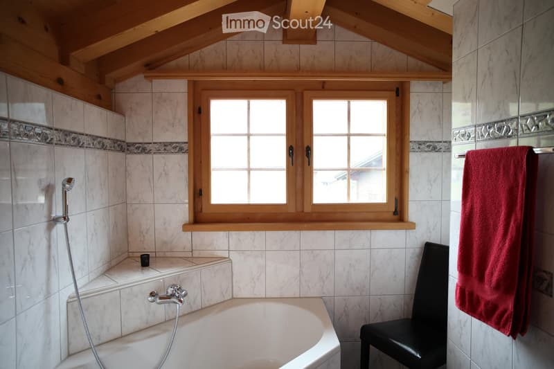 Bathroom with marble tiles, a bathtub, a showerhead, a small stool, and a red towel hanging on the wall. The room features wooden windows and a sloped wooden ceiling.