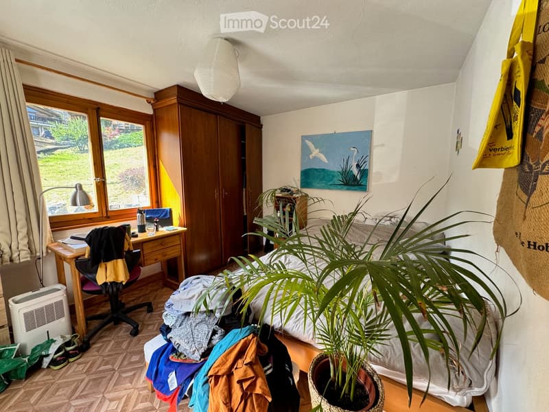 Cozy bedroom with a bed, desk, chair, wardrobe, potted plant, and a painting on the wall. The room features wooden flooring and large windows with curtains, allowing natural light to fill the space.
