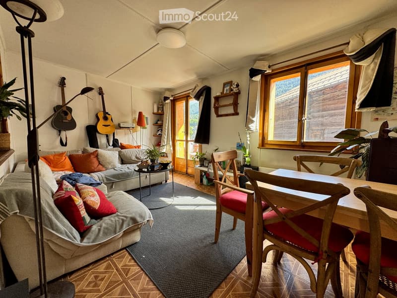 A cozy living room with a grey sofa, wooden dining table, chairs, guitars on the wall, and plants. The room features large windows and a door leading outside.