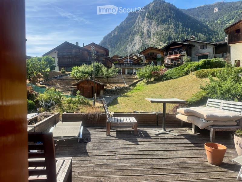 Wooden deck with outdoor furniture including a table, chairs, and a bench, overlooking a scenic mountain view with surrounding houses and greenery.