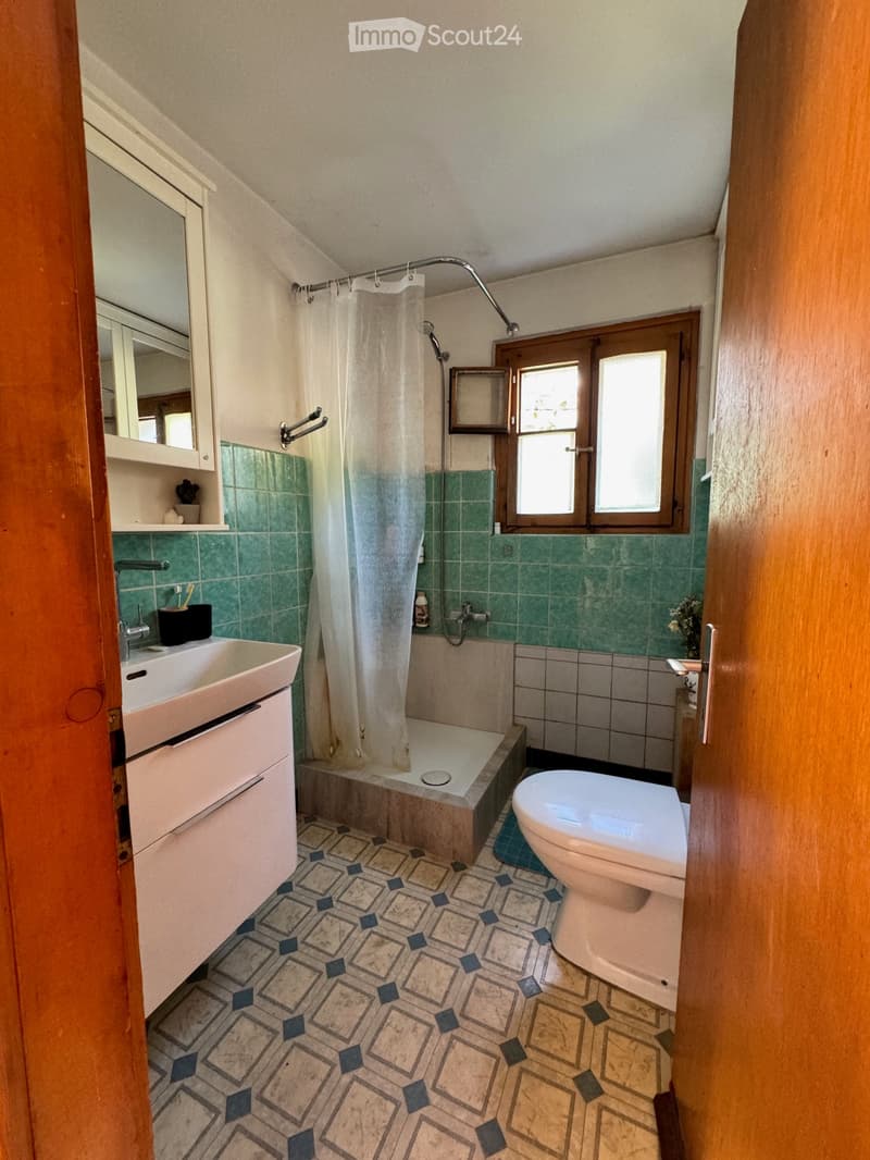 Bathroom with green tiled walls, a shower with a curtain, a sink, a toilet, and a mirror. The floor has a patterned tile design.