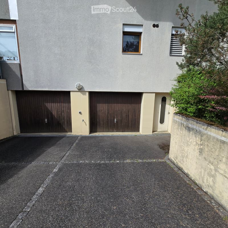 Two-story building with a garage, white facade, and small garden area with shrubs and trees.