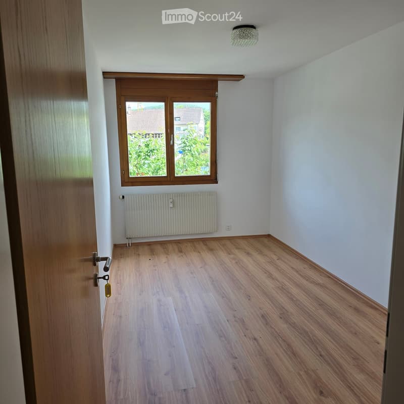 Empty room with wooden flooring, white walls, a window with wooden frame, radiator, and a ceiling light fixture.
