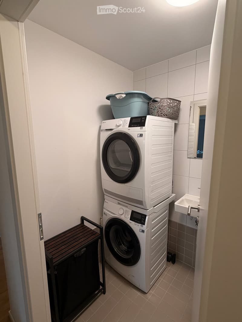 A compact bathroom with a stacked washing machine and dryer, a small sink, a mirror, and a wooden table with a basket on top.
