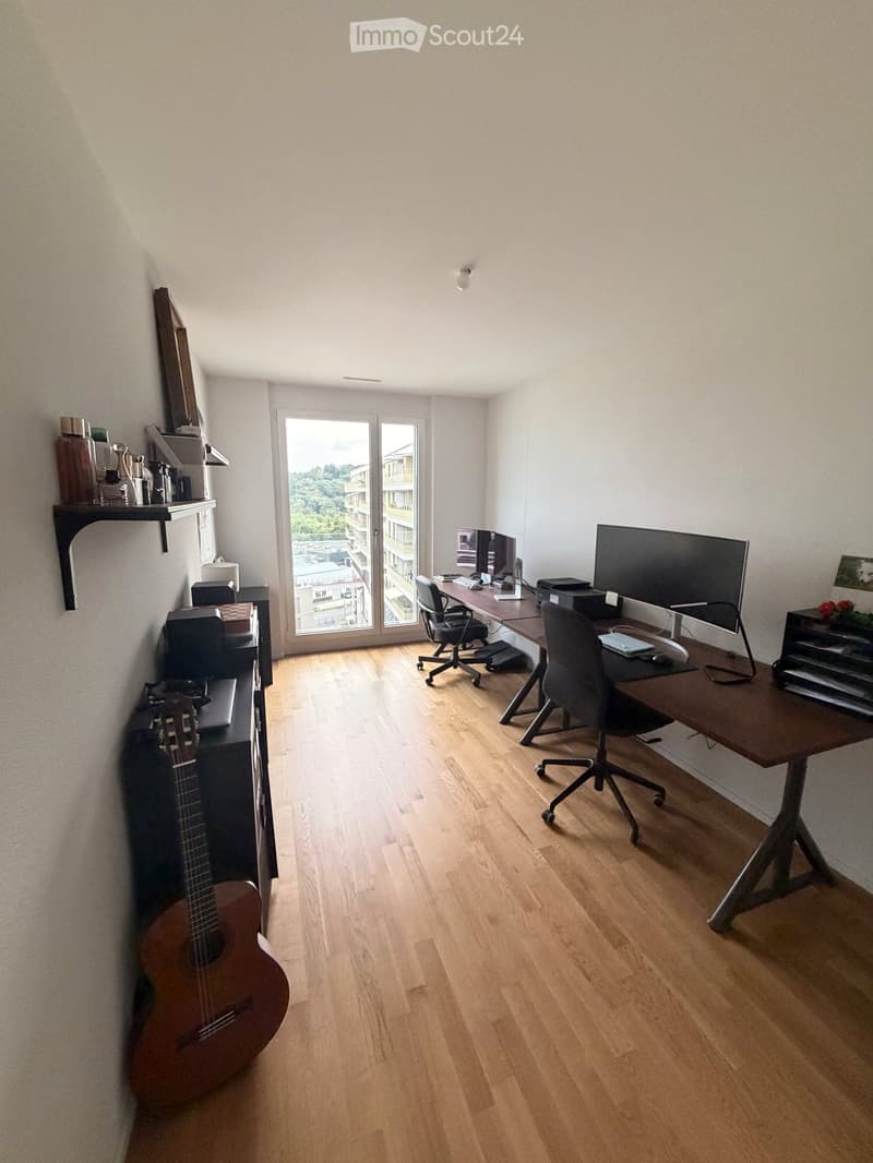 A modern, well-lit room with wooden flooring, a large desk with two chairs, a guitar, and shelves with various items. The room has large windows offering a view of the city.