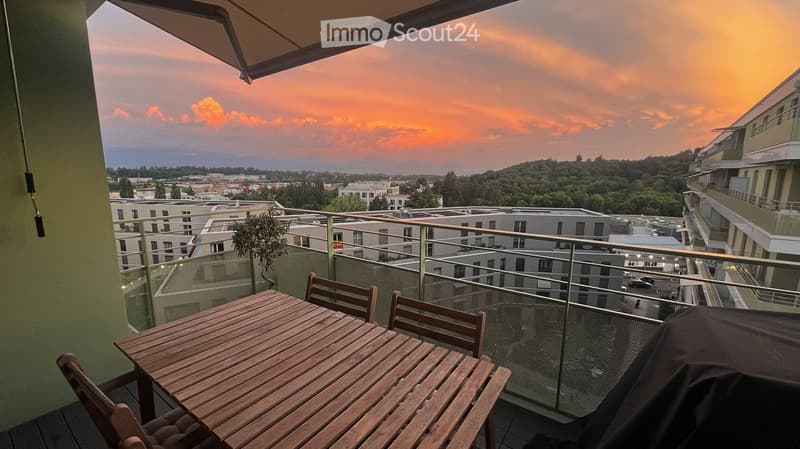 Balcony with a wooden table and chairs, overlooking a cityscape at sunset.