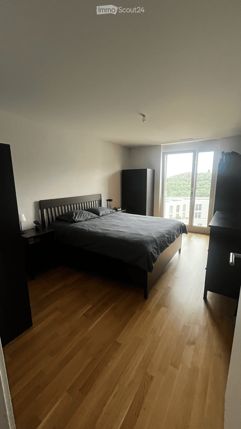 Modern bedroom with wooden flooring, large bed with dark bedding, nightstand with lamp, wardrobe, and large window with view.