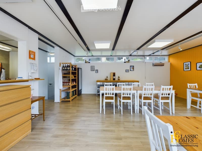 Bright, modern room with wooden flooring, white walls, and slanted ceiling. Features include a long wooden counter, several tables and chairs, a shelf with bottles, and a refrigerator.