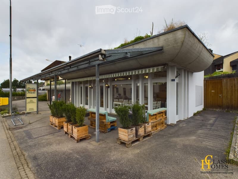 Modern building with a covered outdoor seating area, featuring wooden crates with potted plants, a sign reading "Pasta Hüsti", and a few people visible inside.