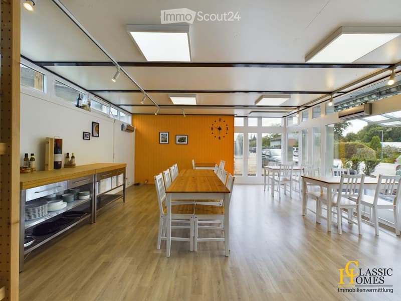 Bright, open-plan room with wooden floors, large windows, and modern lighting. Features a central wooden dining table with chairs, a wooden counter with shelves, and a clock on the wall.