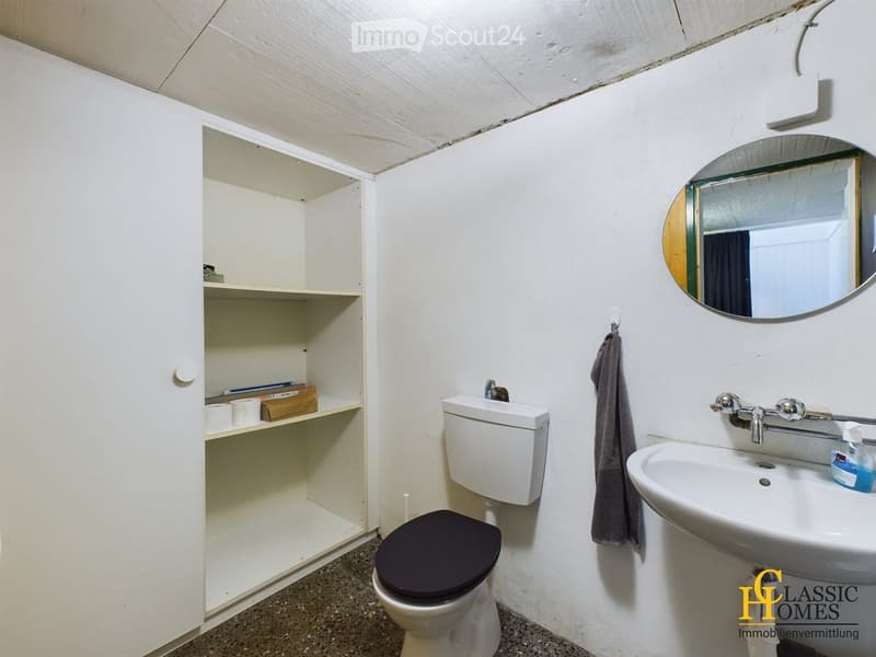 Bathroom with white walls, a toilet, a sink, a mirror, and a cabinet with shelves. A towel is hanging on the wall.