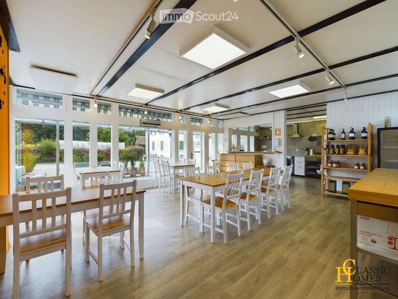 Bright, airy restaurant space with wooden tables and chairs, large windows, and a small kitchen area with shelves and a counter.