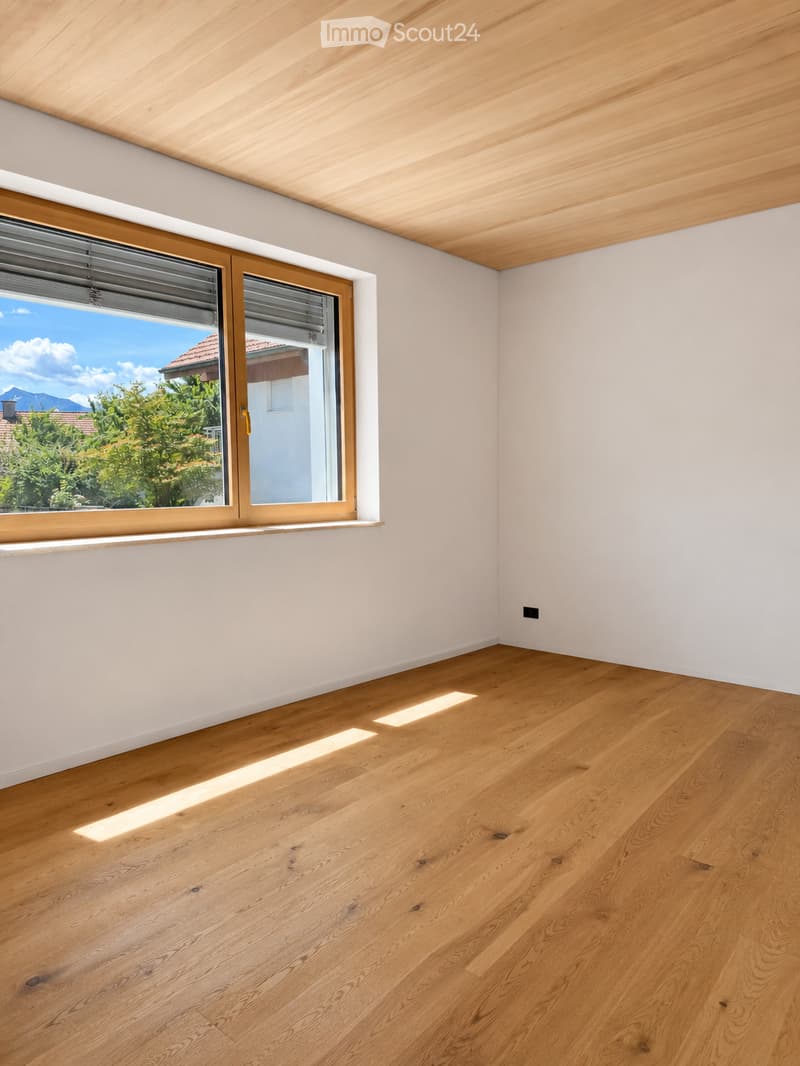 Empty room with light wooden flooring, white walls, and a large window with blinds, allowing natural light to enter.