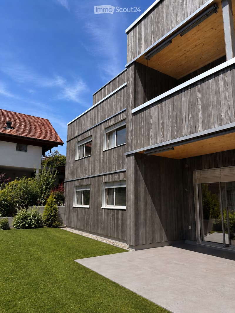 Modern two-story house with wooden siding, large windows, and a covered entrance. The house features a well-maintained lawn and a paved patio area.
