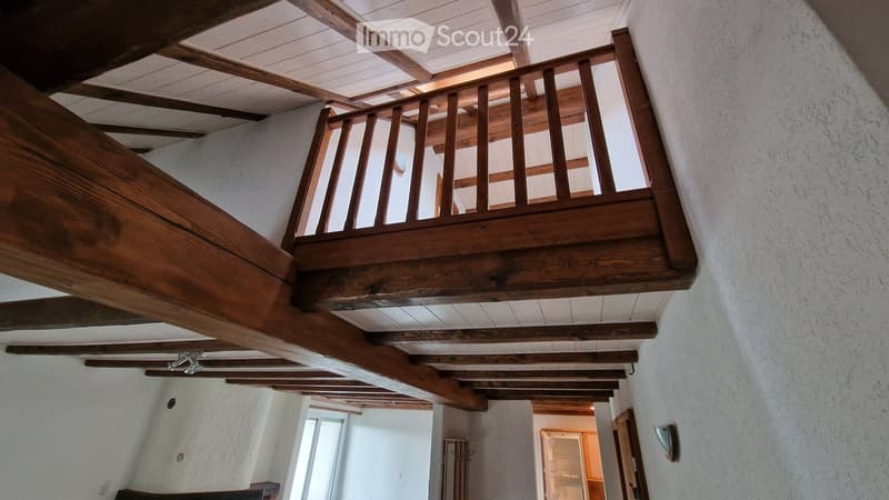 Wooden staircase with a wooden railing and exposed wooden beams on the ceiling. The walls are white, and there is a small window on the left side.