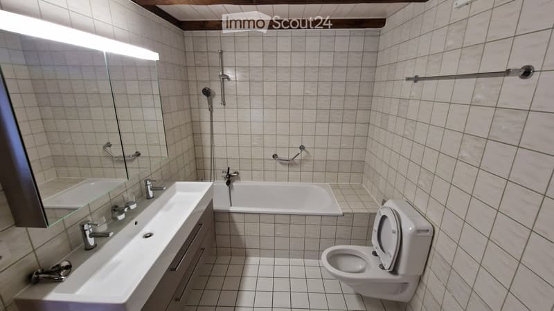 Modern bathroom with white tiled walls and floor, featuring a double sink, bathtub, toilet, and a large mirror with built-in lighting.
