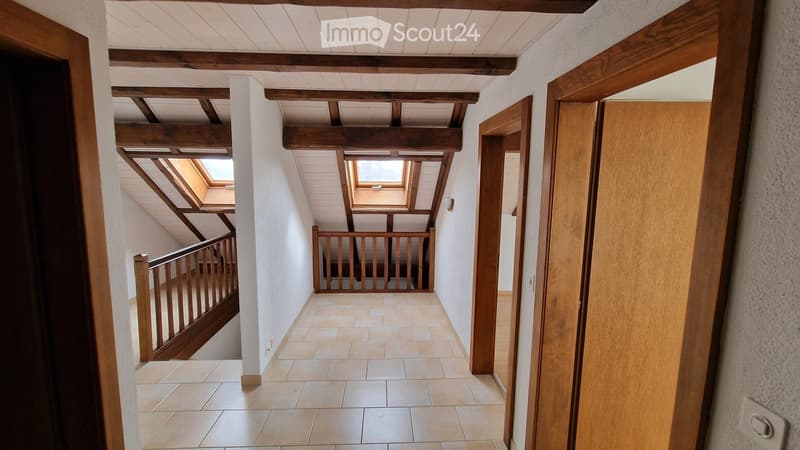 A narrow hallway with wooden beams on the ceiling, tiled floor, and open wooden doors leading to other rooms. The hallway features a staircase with a wooden railing and skylights above.