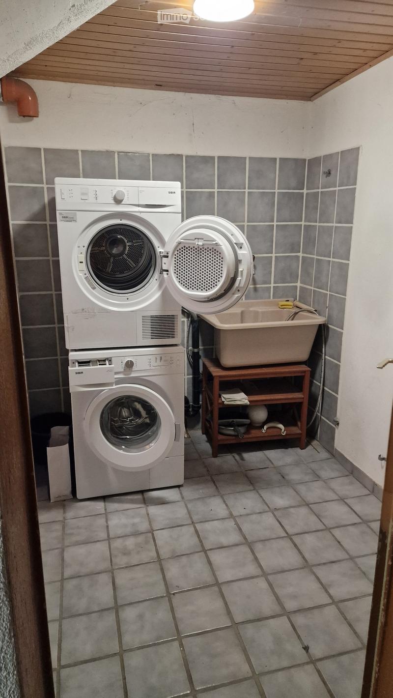 Small bathroom with tiled walls and floor, featuring a stacked washing machine and dryer, a sink on a wooden shelf, and a small wooden shelf holding folded towels and other items.